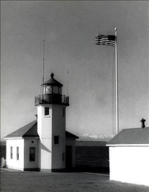 A small lighthouse that is just barely taller than one story stands next to a flag pole flying an American flag. In the distance are snow covered peaks. 